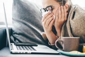 Woman wrapped in a blanket blowing her nose beside a laptop and a mug of tea