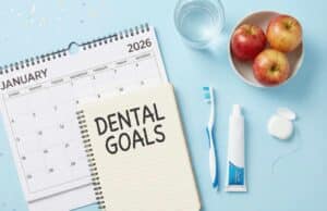Flat lay of dental goals notebook with January 2026 calendar, toothbrush, fluoride toothpaste, floss, apples, and glass of water on light blue background